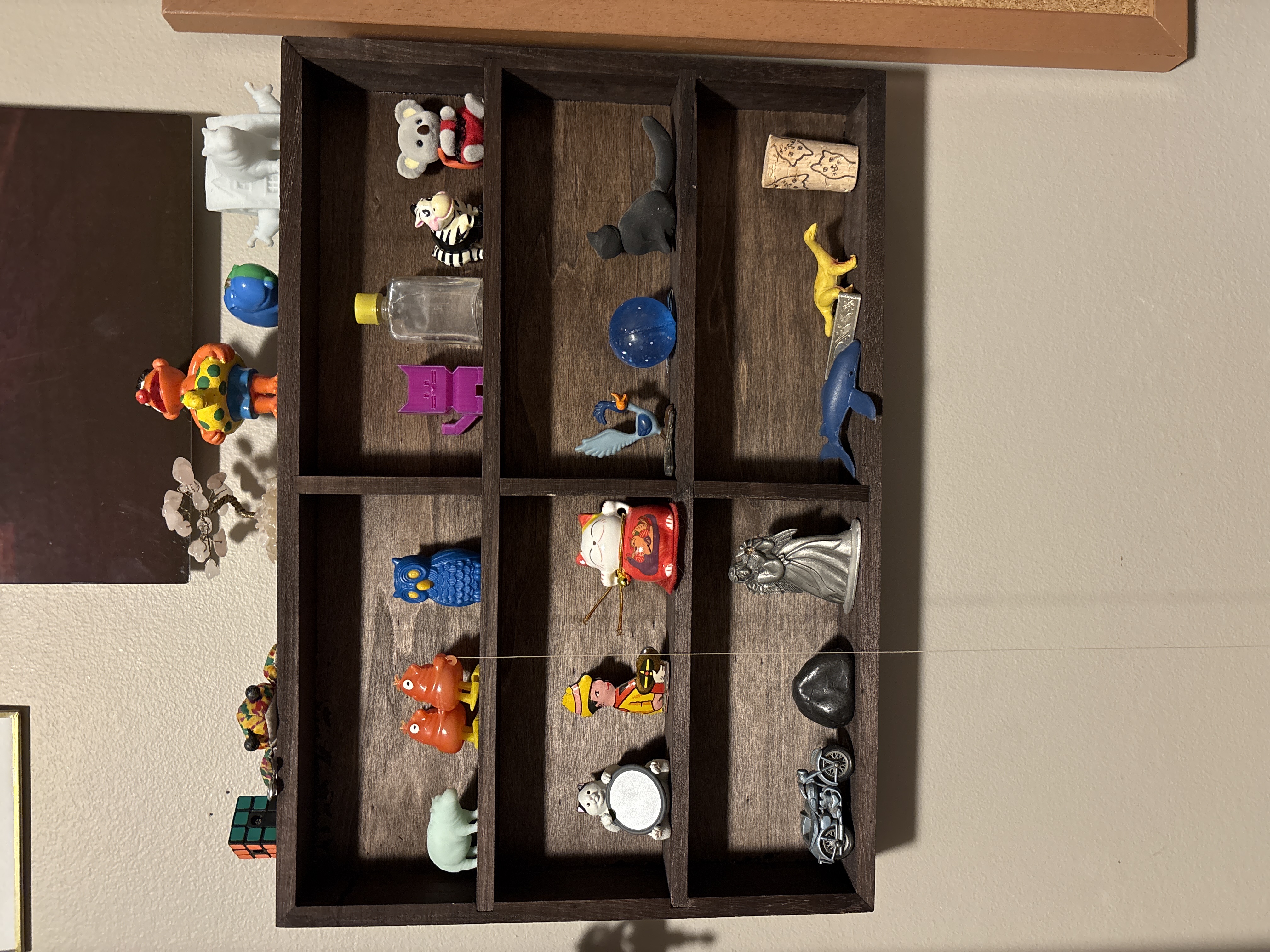 A dark brown shelf with 6 shallow compartments. Each shelf is filled with a variety of “silly little guys” including but not limited to gemstone trees, calico critters, small bottles, and cat figurines.